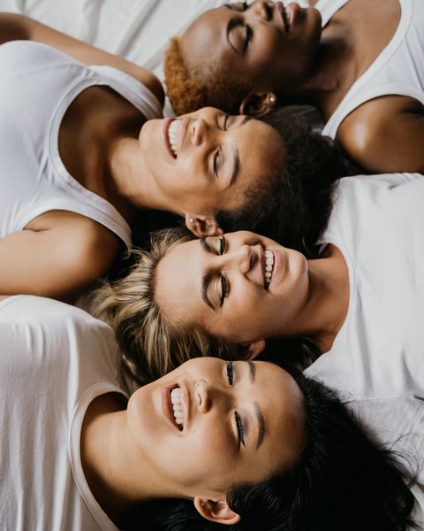Four women in white tops lie with their heads close together on white linen, eyes closed, laughing.