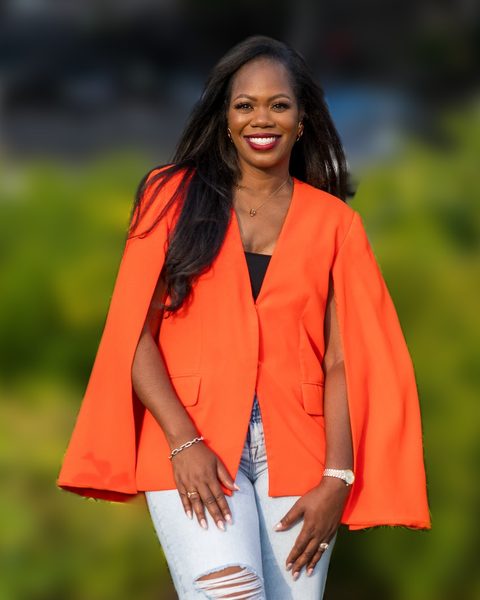 Chericka Johnson, smiling, in an orange cape blazer and light jeans, standing in front of a planted garden bed.