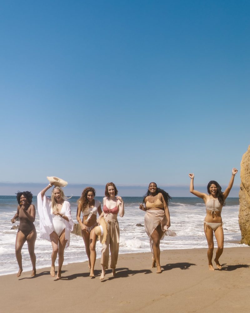 Six women in swimwear and light wraps run laughing toward the camera along a sunny beach, one waving a straw hat and another with both arms raised, a wide blue sky above them.