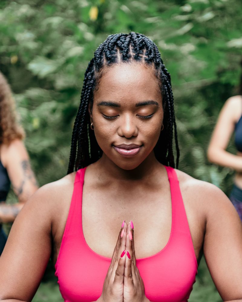 A woman with long braids stands outdoors among green trees, eyes closed and smiling softly, palms pressed together at her chest.